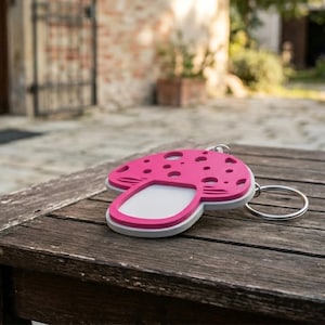 May include: A pink mushroom-shaped keychain with a white and gray border. The keychain has a silver metal ring and is resting on a wooden surface. The mushroom top has several small, round cutouts.