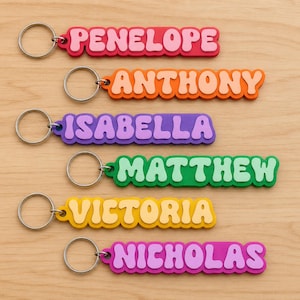 Puede incluir: Una colección de llaveros con nombres coloridos. Cada llavero presenta un nombre diferente: Penelope, Anthony, Isabella, Matthew, Victoria y Nicholas. Los nombres están en un estilo de letras de burbujas, con un borde contrastante y unidos a un llavero plateado.