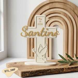 May include: A cream-colored cross with the name "Santino" in gold script. The cross features a dove carrying an olive branch and the date "May 16 2026". The cross is on a round base. A wooden rainbow and cutting board are in the background.