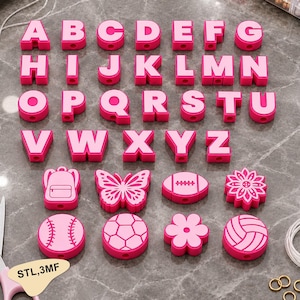 Peut inclure: Une collection de perles en silicone rose et rose clair représentant l'alphabet complet de A à Z, ainsi que des formes incluant un sac à dos, un papillon, un ballon de football, des fleurs, une balle de baseball, un ballon de foot et un volley.