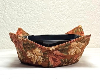 May include: A bowl cosy made from brown fabric, patterned with floral leaves, holding a black rectangular dish. The cosy has a fall colour scheme with brown, orange, and green hues. It is designed to protect hands from hot dishes.