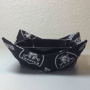 Op de afbeelding: Zwarte en witte komcosy met het Oakland Raiders logo. De cosy is gemaakt van stof en heeft een zwarte kom erin.