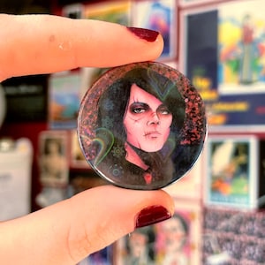 May include: A circular badge with a portrait of a person with dark hair and pale skin, with a scar on their face. The badge has a dark background with red and green accents. The badge is held between two fingers.