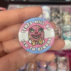 May include: A circular badge with a pink, blue, and white design. It features a cartoon clown face and the text "TERFS WILL BE FED TO BLOBBY". The background incorporates the colours of the transgender pride flag.