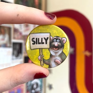 May include: A circular, yellow badge featuring the word "SILLY" in black. A cartoon rat holds a sign with the word "SILLY". The badge is held between fingers with red nail varnish.