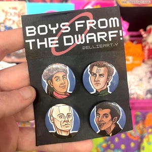 May include: A black card displaying four circular pin badges with character illustrations from "Boys From The Dwarf!". The card reads "BOYS FROM THE DWARF!" in white text with a red oval design. The badges have blue backgrounds.