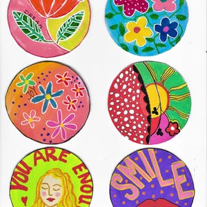 May include: Six circular painted coasters featuring colourful designs. One shows a red flower with the word DREAM, another has blue, pink, and yellow flowers. Others display a floral pattern, a sun motif, a person with the text YOU ARE ENOUGH, and a mouth with SMILE.