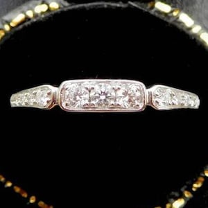 K&ouml;nnte beinhalten: Ein silberner Ring mit einem rechteckigen Mittelstein, flankiert von kleineren Steinen, in einer schwarzen Samt-Schmuckschatulle mit goldenem Rand. Das Ringdesign zeigt eine Reihe kleiner, klarer Edelsteine.