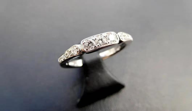 K&ouml;nnte beinhalten: Ein silberner Ring mit drei zentralen Diamanten und kleineren Diamanten an den Seiten. Der Ring ist vor einem dunklen Hintergrund platziert, der sein Funkeln hervorhebt. Ein klassisches Schmuckst&uuml;ck.