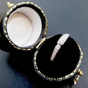 K&ouml;nnte beinhalten: Ein silberner Ring mit kleinen, klaren Steinen wird in einer offenen, schwarzen, runden Schmuckschatulle pr&auml;sentiert. Der Ring liegt auf einem schwarzen Samtinnenraum, und die Schatulle hat goldfarbene Verzierungen und Akzente.