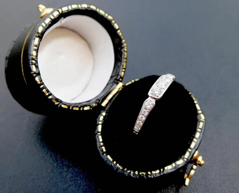 K&ouml;nnte beinhalten: Ein silberner Ring mit kleinen, klaren Steinen wird in einer offenen, schwarzen, runden Schmuckschatulle pr&auml;sentiert. Der Ring liegt auf einem schwarzen Samtinnenraum, und die Schatulle hat goldfarbene Verzierungen und Akzente.