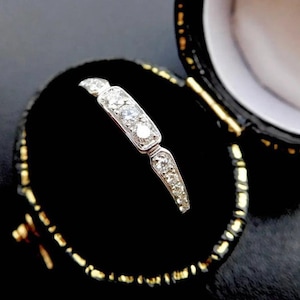 K&ouml;nnte beinhalten: Ein filigraner, silberfarbener Ring mit einem rechteckigen Mittelteil, der mit drei klaren Edelsteinen besetzt ist. Der Ring wird in einer schwarzen Samt-Schmuckschatulle mit goldenen Verzierungen pr&auml;sentiert. Das Design ist elegant und klassisch.