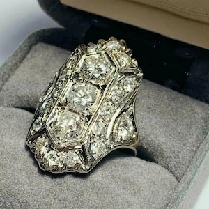 Puede incluir: Un anillo de estilo vintage con una cara rectangular, incrustado con numerosas piedras preciosas claras y brillantes. El anillo presenta una fila central de tres piedras más grandes, rodeadas de gemas más pequeñas en un diseño Art Déco ornamentado. El anillo está engastado en metal plateado.