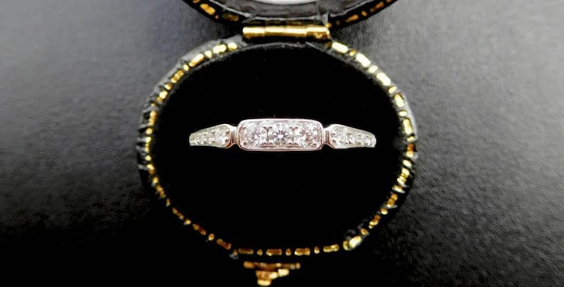 K&ouml;nnte beinhalten: Ein silberner Ring mit einem rechteckigen Mittelstein, flankiert von kleineren Steinen, in einer schwarzen Samt-Schmuckschatulle mit goldenem Rand. Das Ringdesign zeigt eine Reihe kleiner, klarer Edelsteine.