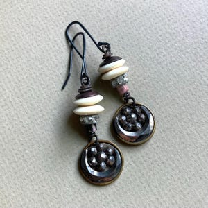 May include: Dangle earrings featuring dark metal hooks, stacked cream-colored disc beads, small grey textured spacers, and a circular pendant base holding a cluster of dark, faceted, metallic-looking beads in a floral arrangement.