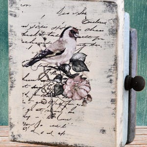 May include: A decorative wooden box with a distressed, vintage aesthetic. The front features a bird perched on a floral branch, with faded script in the background. The box has a dark knob and a light blue side.