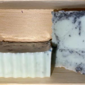 May include: Two handmade soaps with a light green and dark speckled design. One soap has a ridged edge, and the other has a wavy edge. The soaps are displayed in a wooden box.