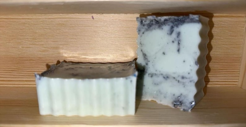 May include: Two handmade soaps with a light green and dark speckled design. One soap has a ridged edge, and the other has a wavy edge. The soaps are displayed in a wooden box.