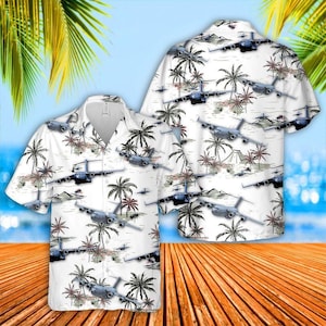 May include: White short-sleeve button-down shirt with a tropical design. The shirt features a pattern of gray airplanes, palm trees, and small islands. The background shows a blue sky and ocean.