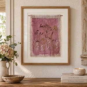 May include: A framed textile art piece featuring a rectangular piece of mauve fabric with frayed edges, suspended from a thin rod. The fabric displays embroidered botanical illustrations of wildflowers in dark brown and gold thread colors.