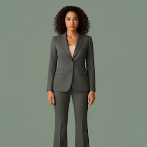 Può includere: Un completo da lavoro grigio antracite composto da un blazer a un bottone con due tasche frontali e pantaloni a zampa abbinati. L'abito è indossato sopra un top rosa chiaro. Lo sfondo è verde tenue.