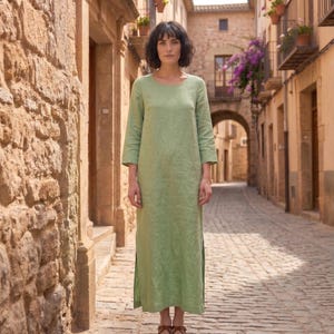 May include: A light green linen dress with three-quarter sleeves and a floor-length hem. The dress has a simple, elegant design and is worn by a person standing on a cobblestone street. The background features stone buildings and an arched doorway.