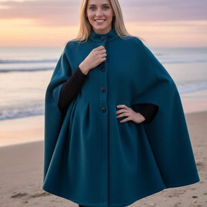 May include: A teal cape coat with a button-up front and a high collar. The coat has a flared silhouette and is worn over a black long-sleeved top and black pants. The background shows a beach with the ocean and a sunset.