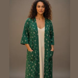 Puede incluir: Una bata tipo kimono verde con estampado floral y bolsillos, usada sobre un vestido blanco. La bata tiene un frente suelto y abierto y mangas tres cuartos. El estampado floral es de color beige claro. La mujer está de pie frente a un fondo neutro.