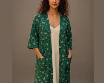 May include: A green floral print kimono-style robe with pockets, worn over a white dress. The robe has a loose, open front and three-quarter sleeves. The floral pattern is a light beige color. The woman is standing in front of a neutral background.