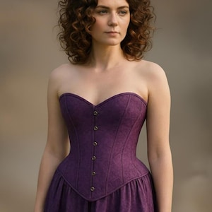 Puede incluir: Un corsé morado oscuro sin tirantes con escote corazón y falda a juego. El corsé presenta una serie de pequeños botones dorados en la parte delantera y detalles sutiles. La prenda está hecha de una tela texturizada.