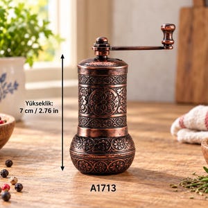 Puede incluir: Un molinillo de especias de metal color cobre con un patrón floral ornamentado y una manivela superior. El molinillo mide 7 cm de altura, como indica el texto Yükseklik: 7 cm / 2.76 in. La base es bulbosa y el cuerpo es cilíndrico.