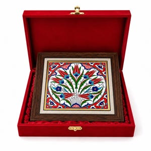 May include: A square ceramic tile featuring a vibrant floral pattern with red tulips and blue accents, set within a dark brown wooden frame. The framed tile is presented inside a rectangular red velvet-lined box with a gold-colored metal latch.