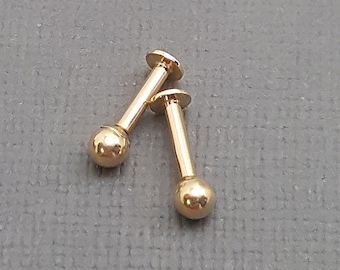 May include: Two gold-colored ball stud earrings. The earrings are made of metal and have a simple, classic design.