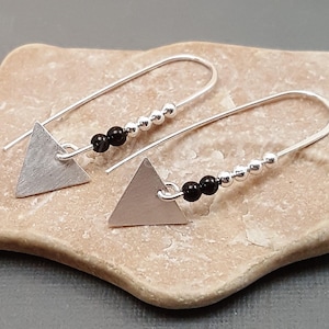 May include: Silver earrings with a geometric design. Each earring has a brushed silver triangle pendant, black beads, and small silver beads on a curved wire. The earrings are displayed on a light brown, textured surface.