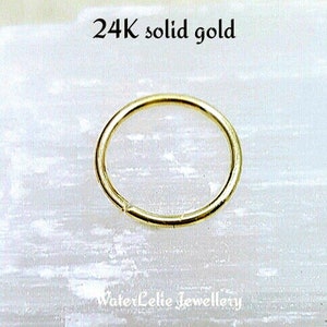 Peut inclure: Gros plan sur une boucle d'oreille créole en or massif 24 carats. La boucle d'oreille est de conception simple et circulaire avec une fermeture invisible. Le texte "24K solid gold" est en haut et "WaterLelie Jewellery" en bas.