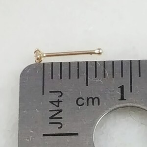 May include: A gold nose ring with a small clear gemstone. The ring is measured against a ruler, showing it is 1 cm long.