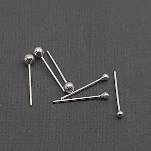 May include: Five silver ball head pins, scattered on a grey surface. The pins are approximately 2.5 cm long.