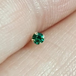 May include: A close-up of a small, green gemstone nose ring. The ring is made of gold and has a small, round gemstone in the centre.