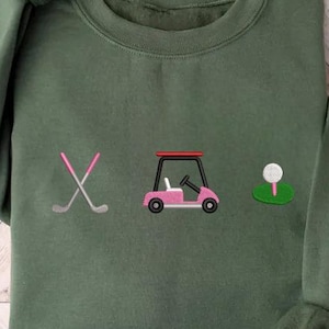 May include: An olive green crewneck sweatshirt featuring an embroidered design of two crossed golf clubs with pink grips, a pink golf cart with a red roof, and a white golf ball resting on a green tee, centered on the chest of the garment.