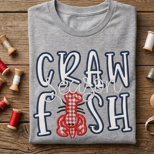 May include: A heather gray t-shirt featuring the text CRAW FISH in bold white letters with dark blue outlines. A red and white gingham patterned lobster graphic is positioned between the words. Several wooden spools of thread surround the shirt.