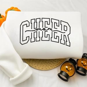 May include: A white sweatshirt featuring the text "CHEER mom" in black, block lettering. The sleeve is folded over. Two orange pumpkins and two decorative lanterns are in the background.