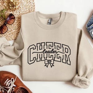 May include: A beige sweatshirt with the word "Cheerleader" in black block letters. A black bow is below the word "leader". The sweatshirt is paired with blue jeans and brown shoes.