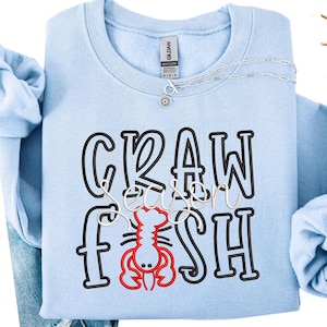 May include: Light blue sweatshirt with the words "CRAWFISH SEASON" in black and white lettering, and a red crawfish graphic. A silver necklace with a small pendant is draped across the top. The sweatshirt is folded.