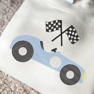 May include: Cream-colored sweatshirt featuring an embroidered light blue race car with black wheels and a white circle. Two crossed checkered flags are above the car. A fun design for kids.