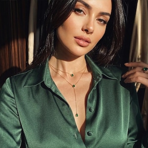 May include: A woman wearing a gold necklace with three green emerald pendants. She is also wearing a matching emerald ring. The woman is wearing a dark green satin button-down shirt.