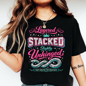 Puede incluir: Camiseta negra con un diseño gráfico que presenta las palabras "Layered, Stacked, Slightly Unhinged" en rosa, plata y turquesa. Un diseño de serpiente está debajo del texto, con las palabras "Team Created for Greatness".
