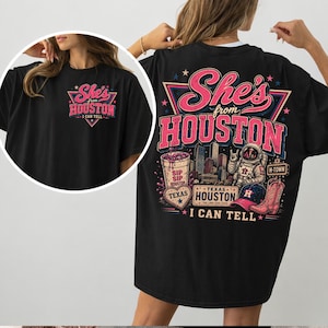 May include: A black t-shirt featuring a graphic design with the text She's from HOUSTON I CAN TELL. The back print includes a Houston skyline, an astronaut, a cowboy boot, a baseball cap, a heart, and a cup with the text SIP SIP HOUSTON.
