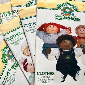 May include: Several vintage Cabbage Patch Kids clothing pattern packets. The packets feature illustrations of Cabbage Patch Kids dolls. The packets are labeled "CLOTHES For your Cabbage Patch Kids!"