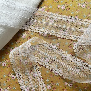 May include: White lace trim with a scalloped edge is displayed on a floral patterned fabric. The lace is a delicate, open weave design with a decorative edge. The fabric has a mustard yellow background with small pink and white flowers.