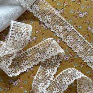 May include: White lace trim with a scalloped edge, displayed on a floral patterned fabric. The lace is intricately designed with a delicate pattern. A folded white fabric is also visible in the image. The background features a mustard-colored fabric with small pink and white floral designs.